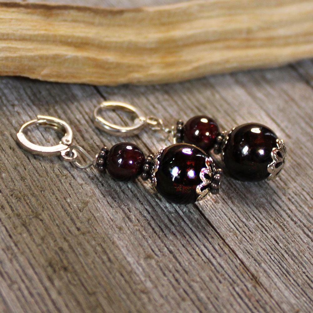 A pair of Sacred Geometry Garnet Earrings featuring glossy dark red beads and sterling silver lever back earwires, artisan handmade and displayed on a rustic wooden surface next to driftwood.