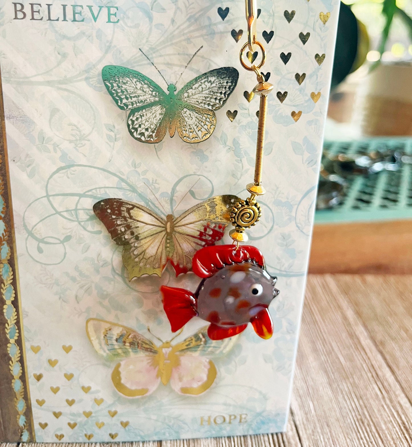 The Gold Metal Bookmark, a unique Glass Fish Bookmark with three butterflies and a dangling glass fish charm, makes a perfect gift for aquarists or a fun aquatic page holder—shown on wood in natural light.