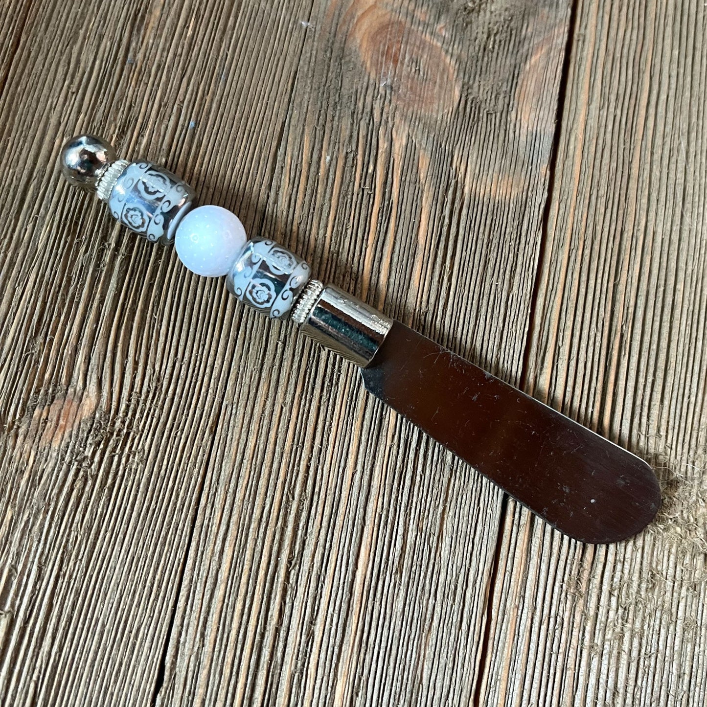 The Handcrafted Golf Ball Dip Knife: Novelty Golf Gift for Charcuterie features a dark blade and a beaded handle with silver and white accents, making it a charming golf-themed kitchen accessory displayed on a textured wooden surface.