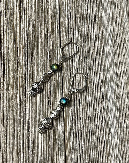 Fish Dangle Earrings by Gwynstone Handmade feature silver-tone fish charms and color-changing mood beads—perfect as an ocean lover gift. Displayed on a textured wooden surface.