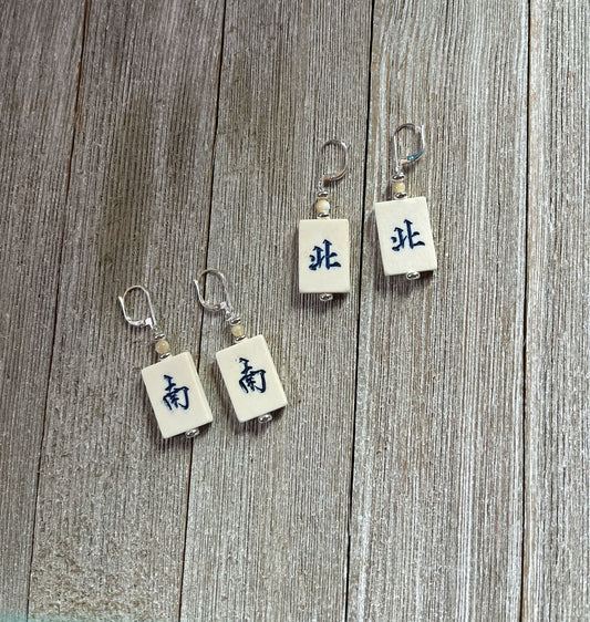 Four Vintage Style Majong Tile Earrings: Handcrafted North/South Winds, each with metal hooks and white tiles featuring black Chinese characters, are displayed on wood. Two earrings share the same wind tile; the other two show different winds.
