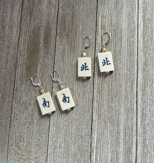 The Mahjong Tile Earrings: North/South Winds, perfect for mahjong players, feature four rectangular tiles with metallic hooks and black Chinese characters, arranged on a weathered wooden surface for a unique, Asian-inspired look.