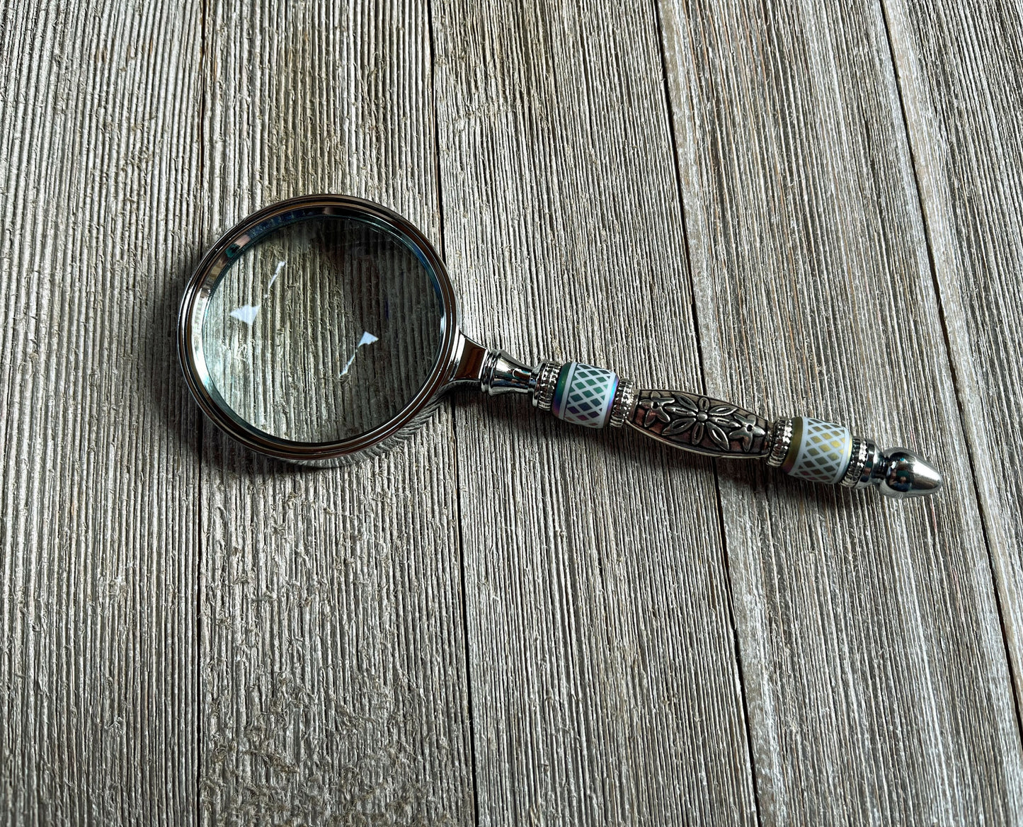 The Shimmering Bead Handed Magnifier for Fine Details, a unique office crafting tool with an ornate handle, rests on textured wood—ideal for jewelry making or intricate beadwork.