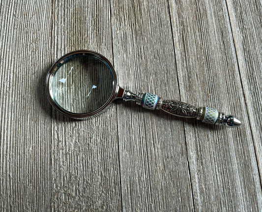 The Shimmering Bead Handed Magnifier for Fine Details, a unique office crafting tool with an ornate handle, rests on textured wood—ideal for jewelry making or intricate beadwork.
