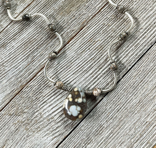 The Ocean Jasper Sterling Silver Jewelry Set features an artisan wave pendant necklace with a wavy design and a sterling silver teardrop pendant adorned with white and yellow flower patterns, displayed on weathered wood.