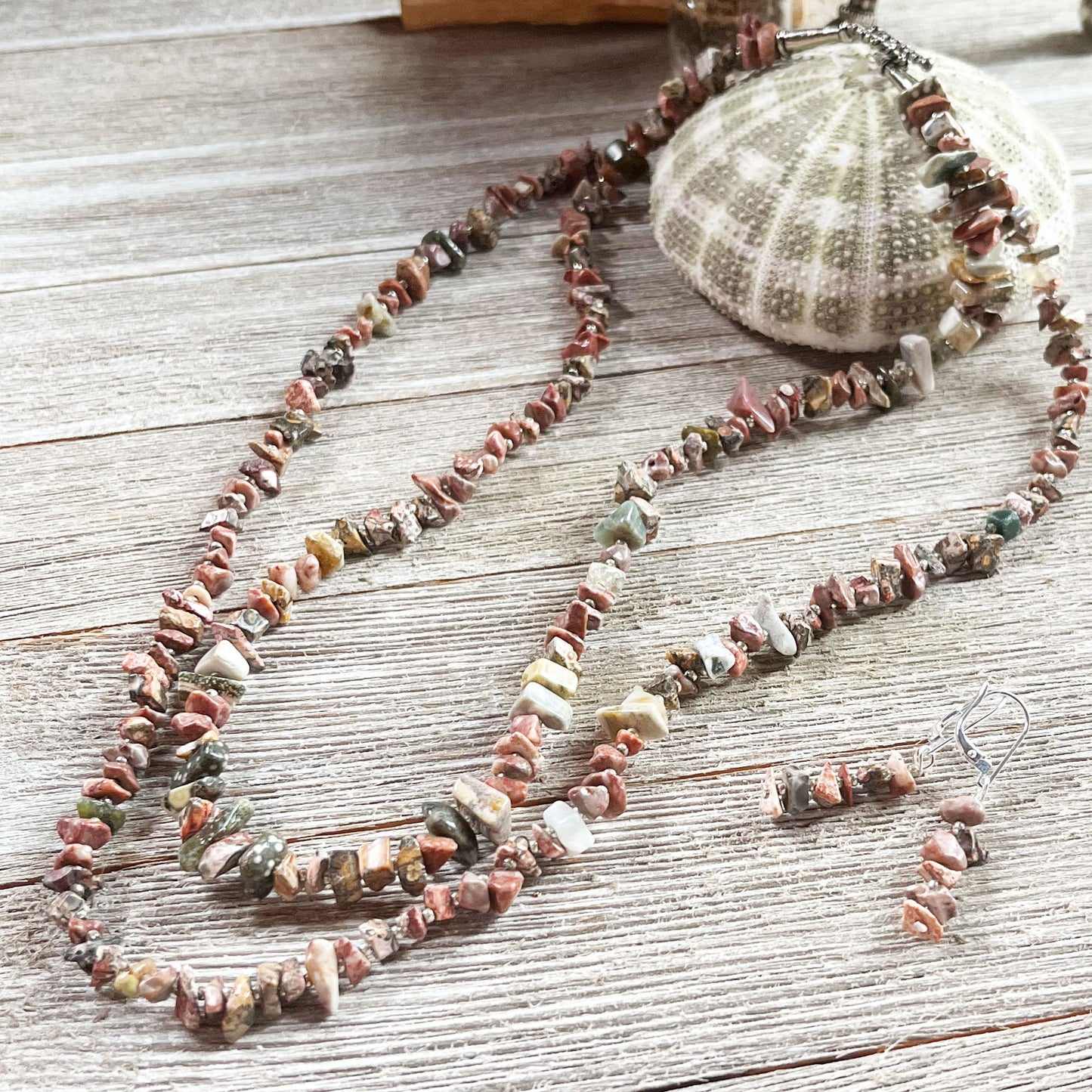 The Jasper Fragment Necklace and Earrings Set—sterling silver beachcomber jewelry featuring irregular, multicolored stone chips—rests on light wood beside a sea urchin shell.