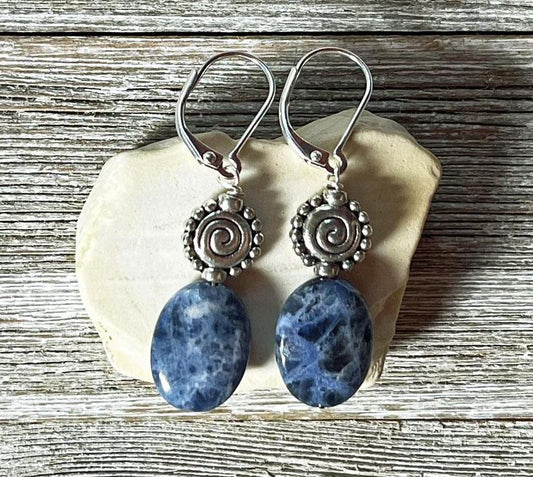 Artisan-designed Sodalite earrings with silver spiral accents, featuring dark blue oval stones, displayed on a pale seashell over rustic wood—unique gemstone jewelry for creativity, intuition, and self-expression.