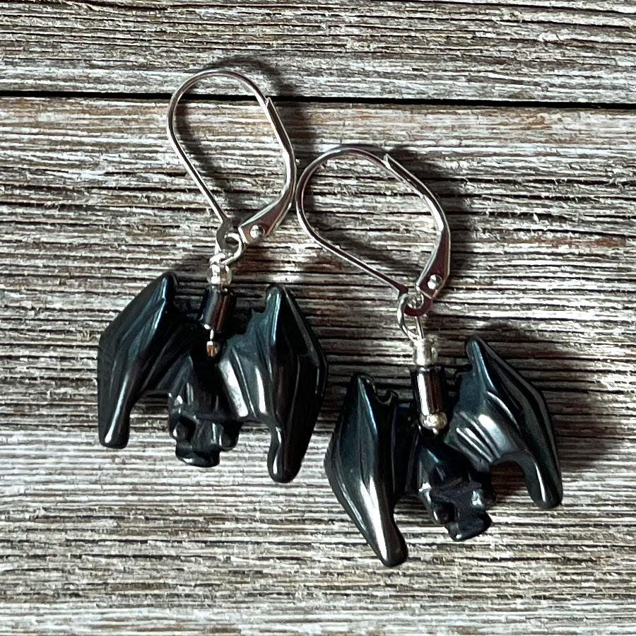 The Spooky Hematite Earrings for Halloween feature small black bat-shaped charms with spread wings and silver hooks, displayed on rustic wood. Perfect as good luck symbol creepy bat jewelry for the Halloween season.