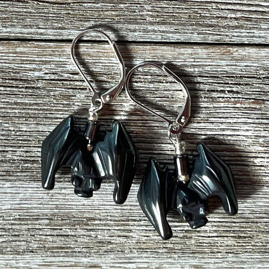 The Spooky Hematite Earrings for Halloween feature small black bat-shaped charms with spread wings and silver hooks, displayed on rustic wood. Perfect as good luck symbol creepy bat jewelry for the Halloween season.