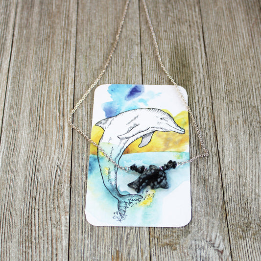 The Handcrafted Snowflake Obsidian Dolphin Necklace: Sea Life Jewelry is shown atop an illustrated card with a dolphin leaping from water, set on rustic wood, showcasing its unique stone beads and ocean-inspired charm.