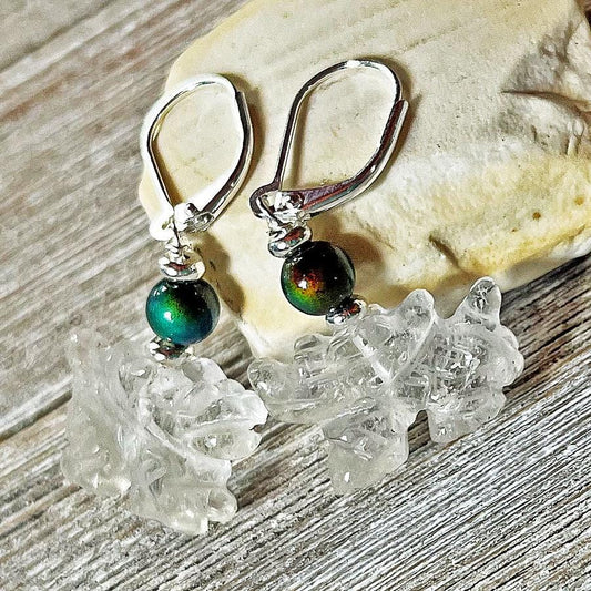 The Clear Dragon Earrings are handmade fantasy jewelry featuring color-changing quartz, iridescent dark green beads, and sterling silver hooks, displayed on rustic wood with a light rock in the background.