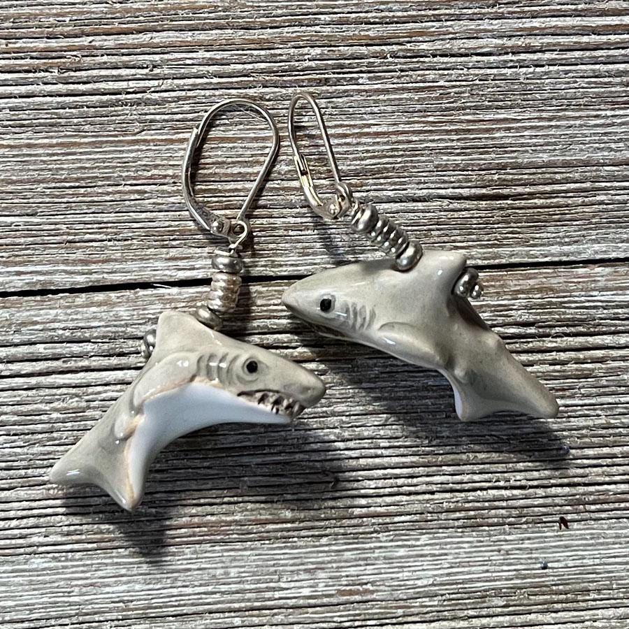 Grinning Shark Earrings: Hand-painted Ceramic Lever Back Jewelry in gray with white undersides, detailed fins, eyes, and teeth. Shown on a weathered wooden surface.