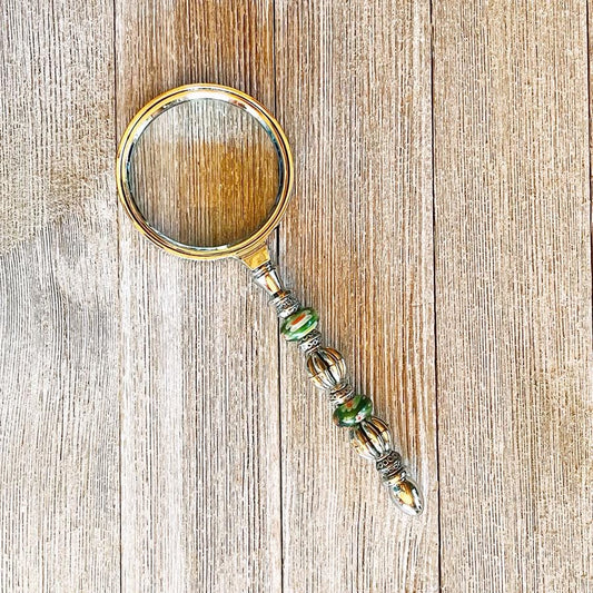 The Daisy Green Beaded Magnifier, a handmade silver office crafting tool with green accents, rests on a wooden surface displaying its unique beaded handle and elegant design.