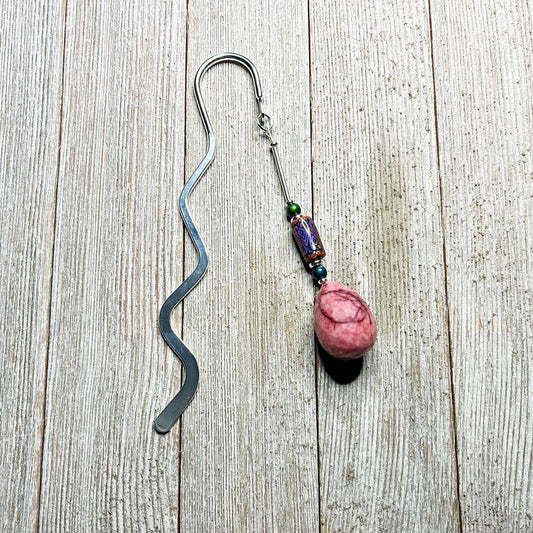 The Handcrafted Stone Bookmark: Teardrop Gemstone, with a wavy silver design and a pink stone charm accented by colorful beads, rests on light wood—a thoughtful gift for book lovers.