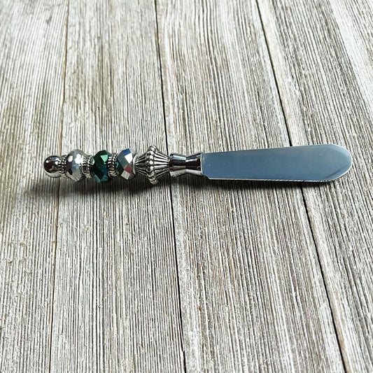 The Dark Faceted Glass Beaded Cheese Knife: Handcrafted Dip Spreader features a faceted glass bead on its metal handle, bringing elegance and style to any table setting when placed on a rustic wooden surface.