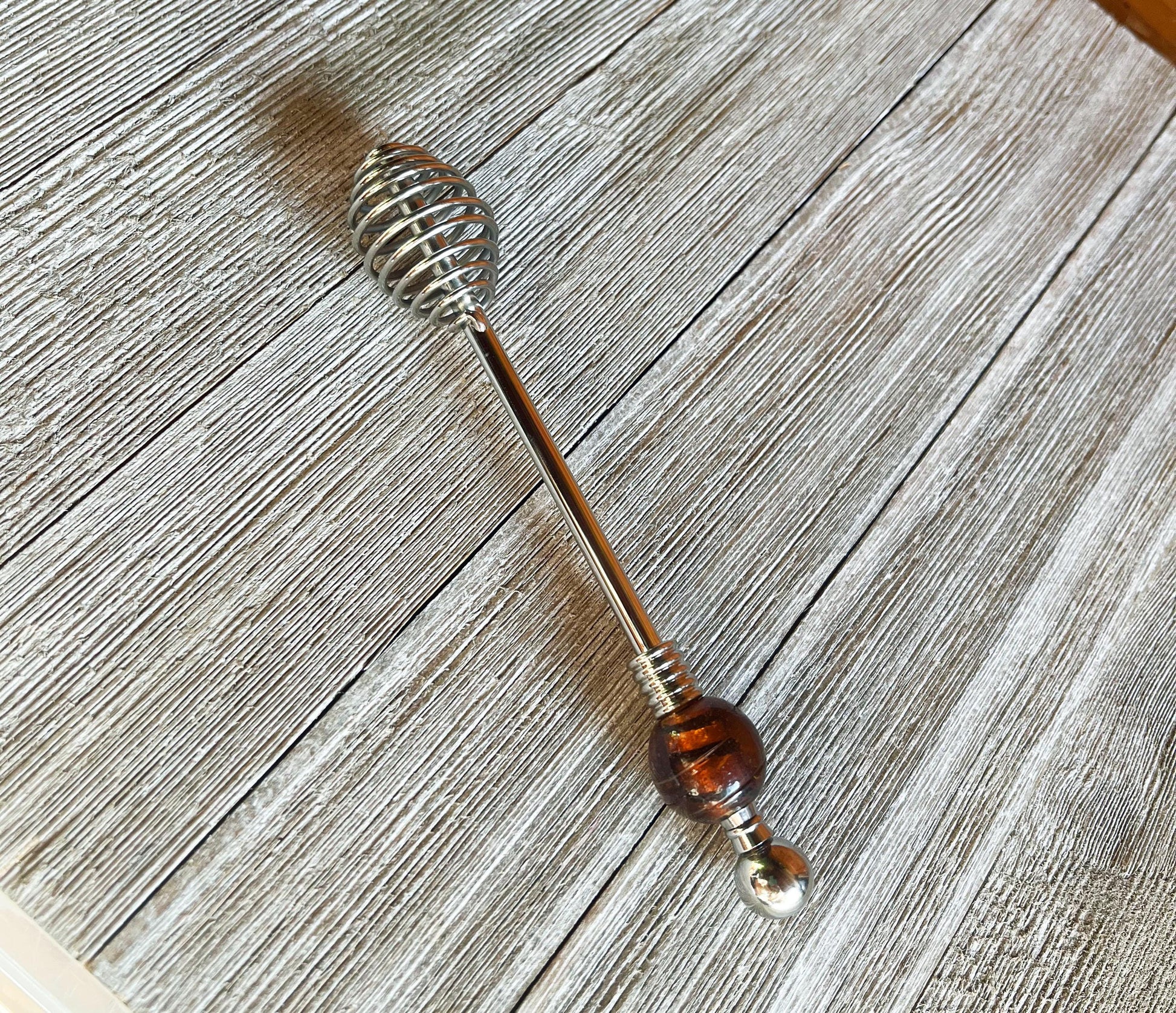 The Artisan Honey Drizzler features a spiral metal end and a brown & golden art glass bead, measuring 6 inches, shown resting on a textured wooden surface.