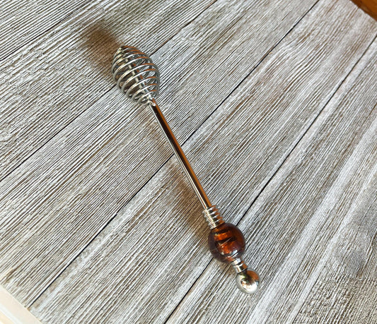 The Artisan Honey Drizzler features a spiral metal end and a brown & golden art glass bead, measuring 6 inches, shown resting on a textured wooden surface.