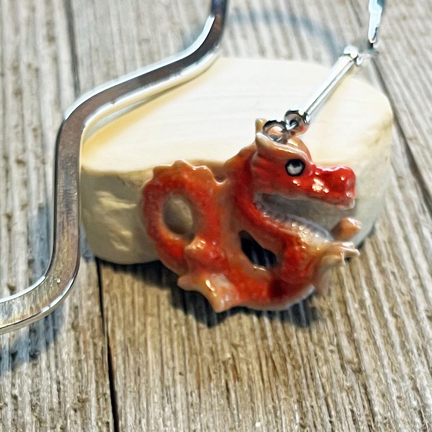 A close-up of the Red Dragon Bookmark—a handmade porcelain fantasy gift—shows its dragon-shaped pendant with a silver hook on a white oval stone, all set atop a textured wooden surface.