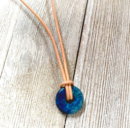The Minimalist Lapis Lazuli Necklace features a blue circle stone pendant on an Italian leather cord, tied in a simple knot and displayed on a weathered wooden surface.