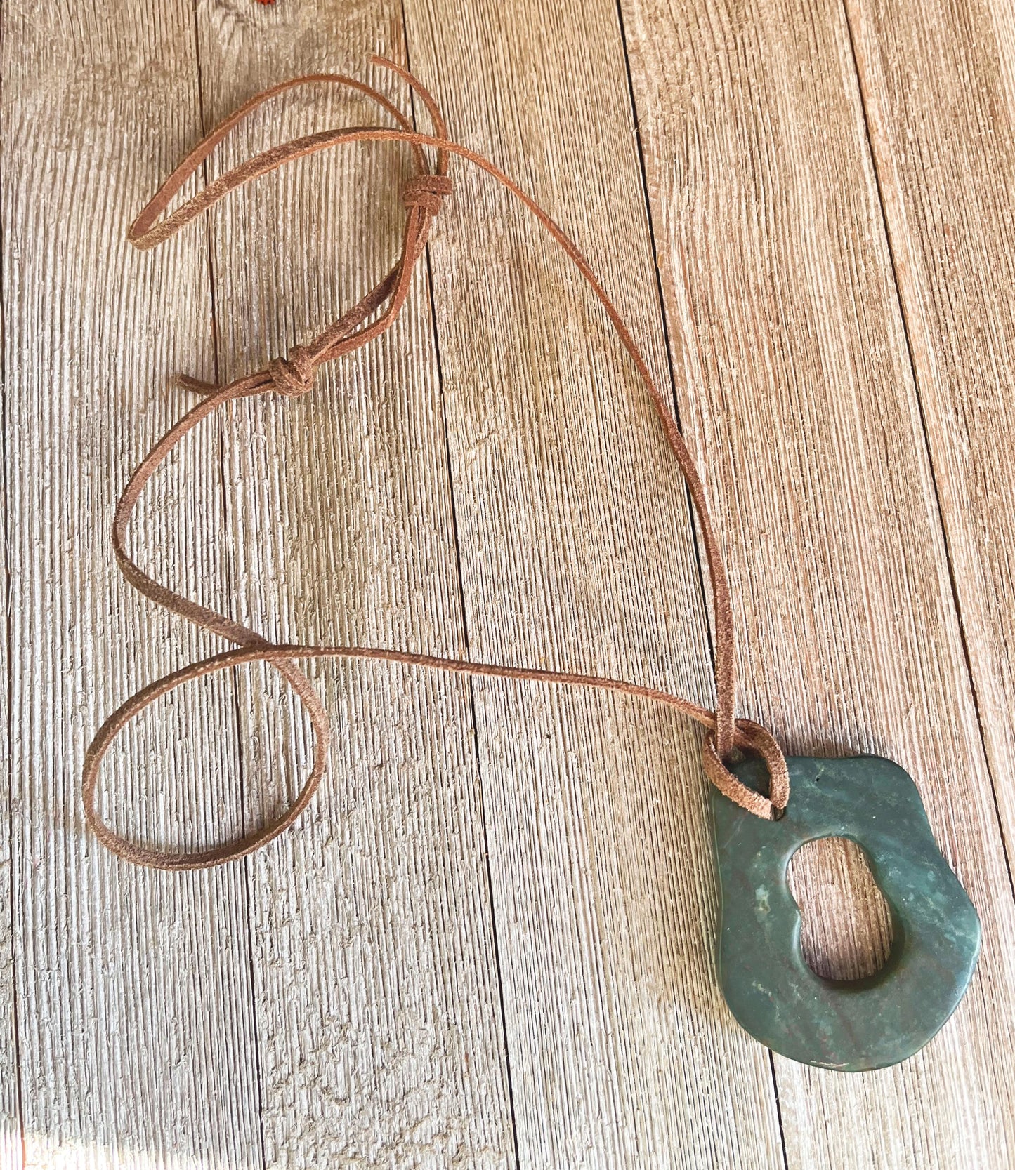The Handcrafted Green Aventurine Pendant Necklace features an irregular greenstone pendant with a central hole on an adjustable leather cord, displayed on a light wooden surface.