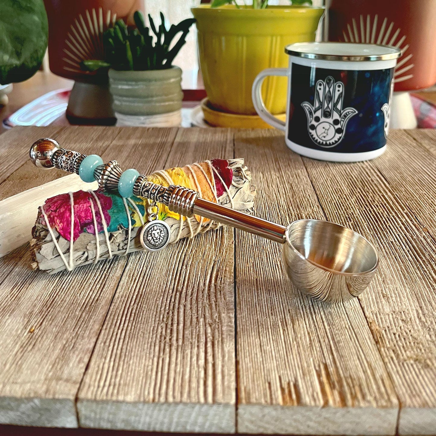 A bundle of sage with colorful strings and beads rests next to the Zodiac Sign Coffee Scoop – Beaded Zen Herb Spoon, Fire Element, on a wooden surface. Behind them are potted plants and a mug featuring a hamsa hand design.