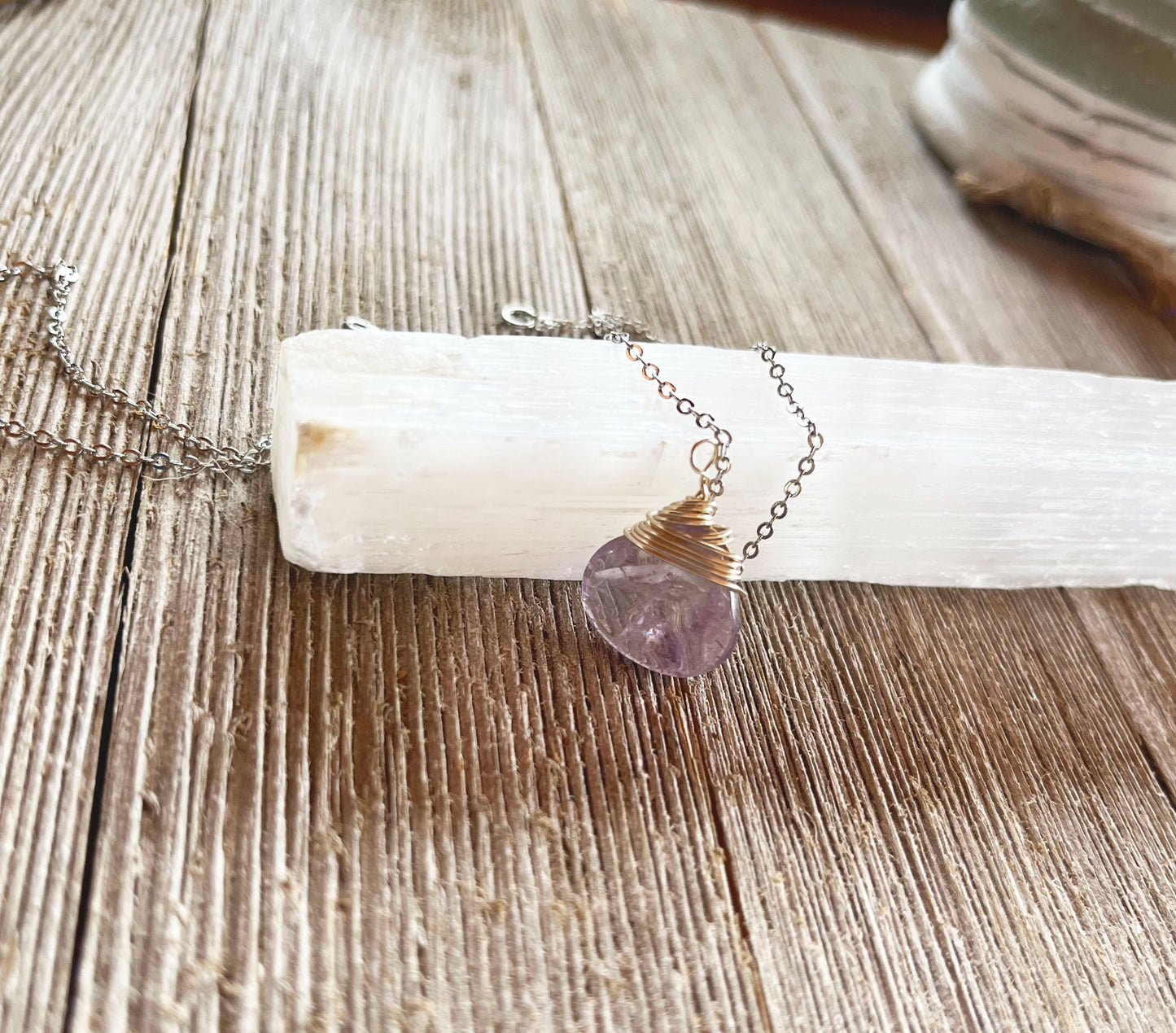 The Wire Wrapped Amethyst Briolette Necklace: Bird's Nest Zen Jewelry features a purple gemstone pendant on a silver chain, wrapped in gold wire and shown atop a white selenite crystal stick on rustic wood—ideal for zen style.