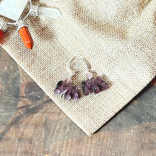 Handcrafted Purple Rhino Earrings with optic stone—perfect wildlife gift—feature purple rhino shapes and silver hooks, displayed on burlap beside a crystal pendant with orange and clear stones atop a rustic wooden surface.