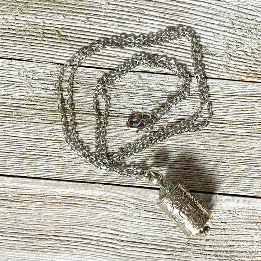 The Prayer Wheel Necklace features a silver Zen Mantra Pendant with intricate patterns, designed for mindful meditation, shown resting on a textured wooden surface.