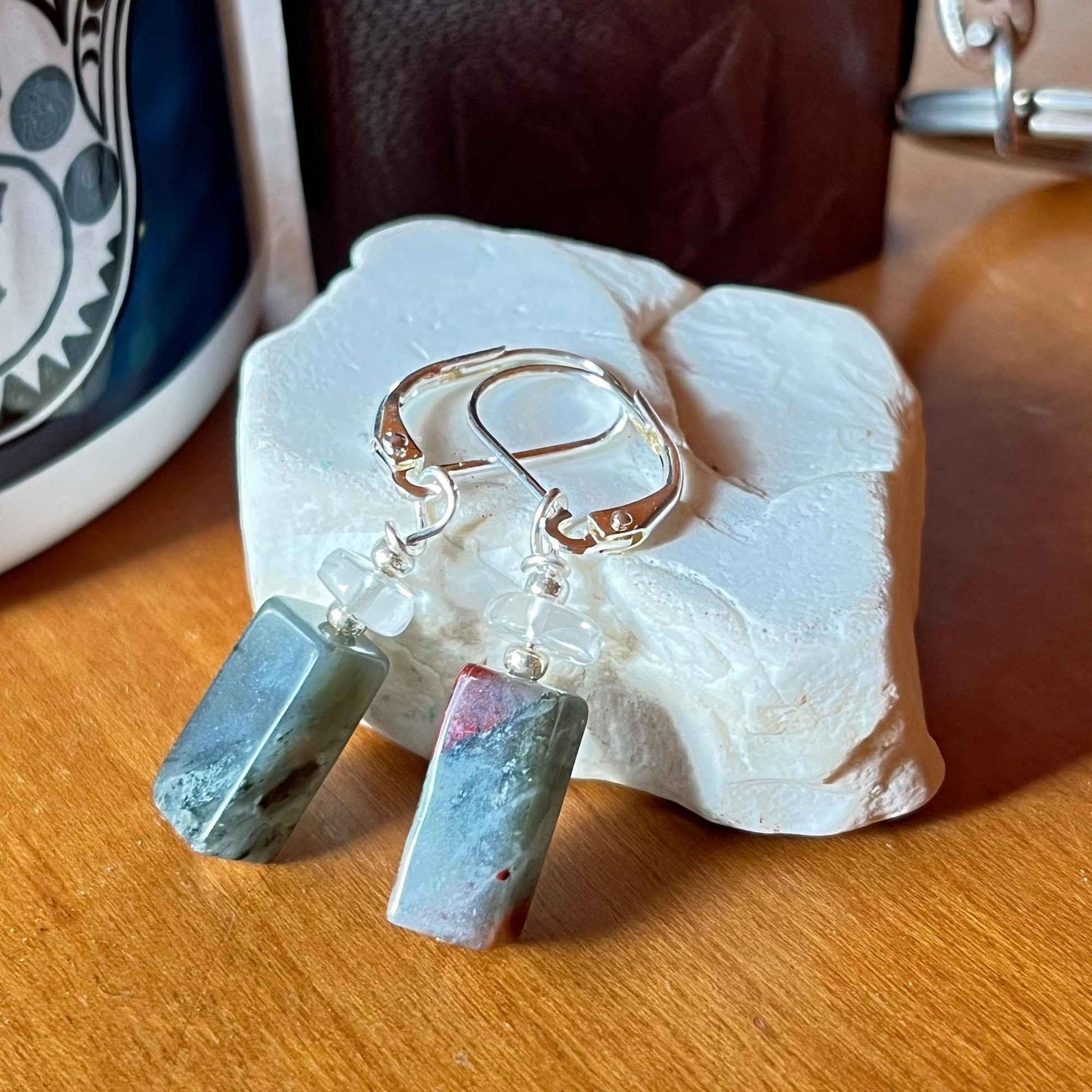 Spiritual Bloodstone Dangle Earrings with Clear Quartz: minimalist Zen jewelry featuring silver hoops, rectangular green and gray stone pendants, and translucent beads, displayed on white textured stone near a ceramic cup.