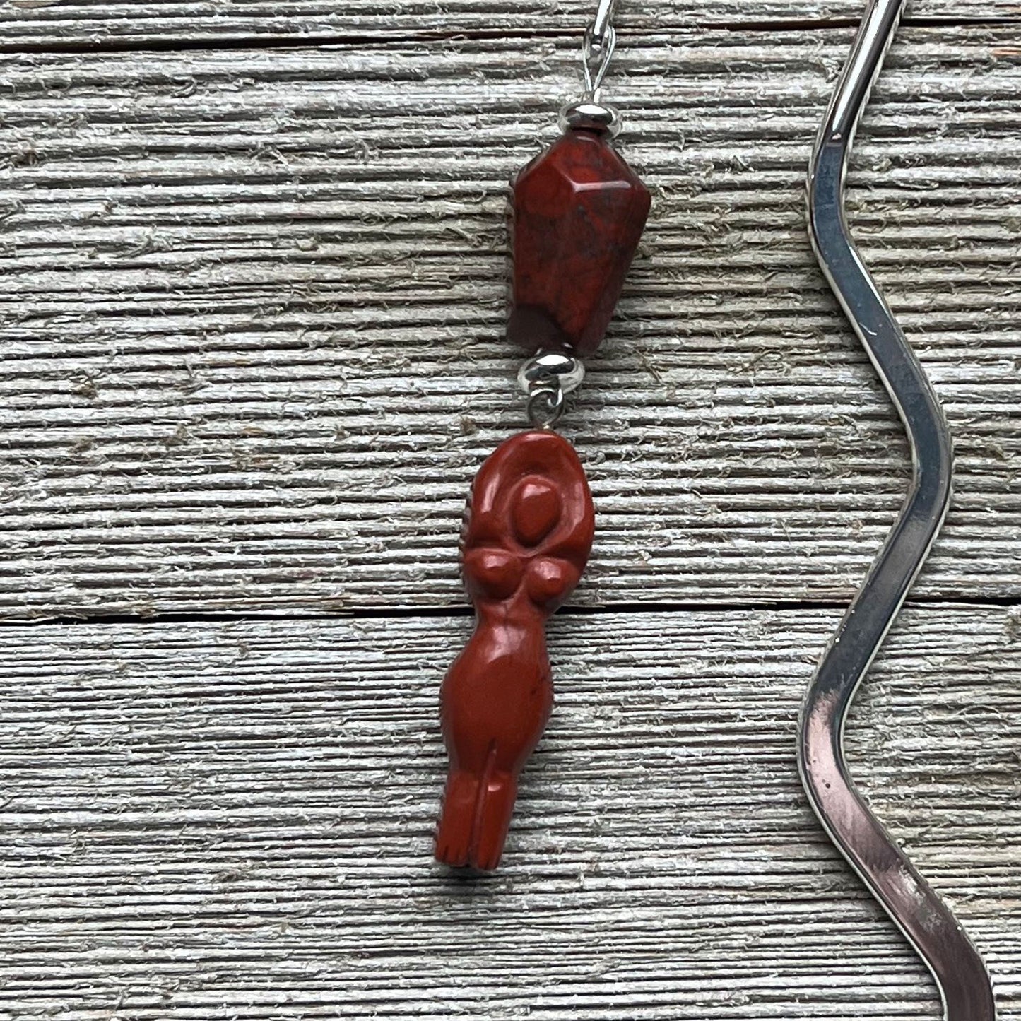 The Handcrafted Red Jasper Goddess Bookmark features a wavy silver handle, a red jasper bead, and a red goddess charm on metal loops—a unique Divine Feminine Gift displayed on a textured wooden surface.