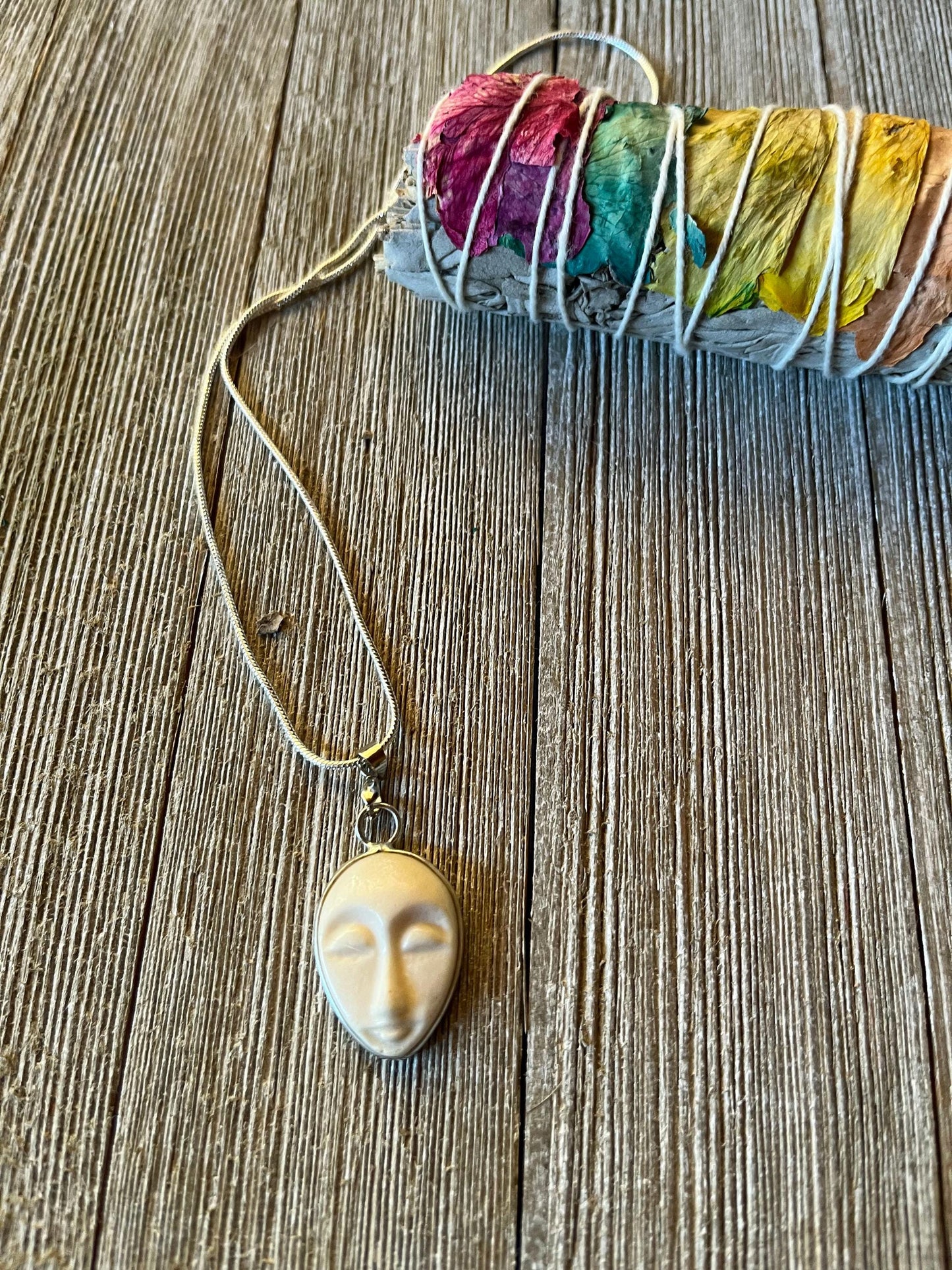 The Serene Carved Bone Face Necklace—Tranquil Talisman for Calm & Clarity—rests beside dried sage and flower petals, offering a unique spiritual accessory to inspire mindfulness and peace.