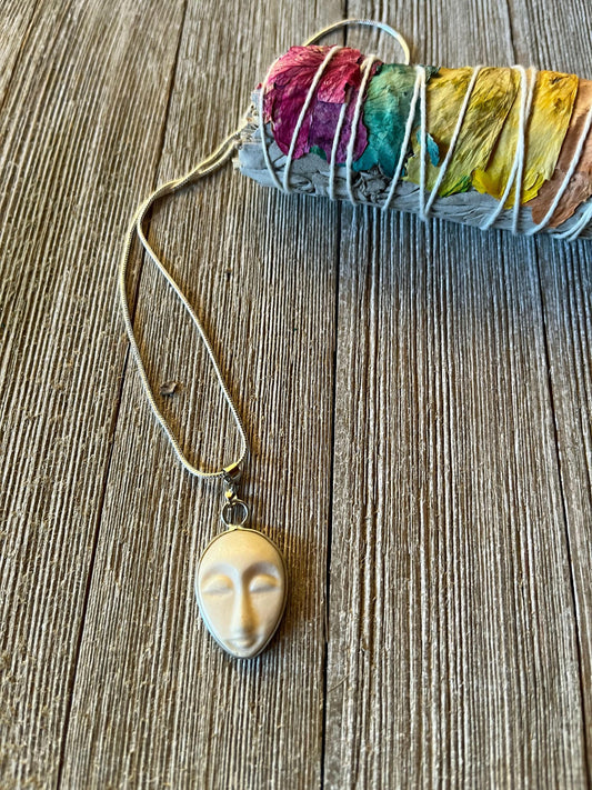 The Serene Carved Bone Face Necklace—Tranquil Talisman for Calm & Clarity—rests beside dried sage and flower petals, offering a unique spiritual accessory to inspire mindfulness and peace.