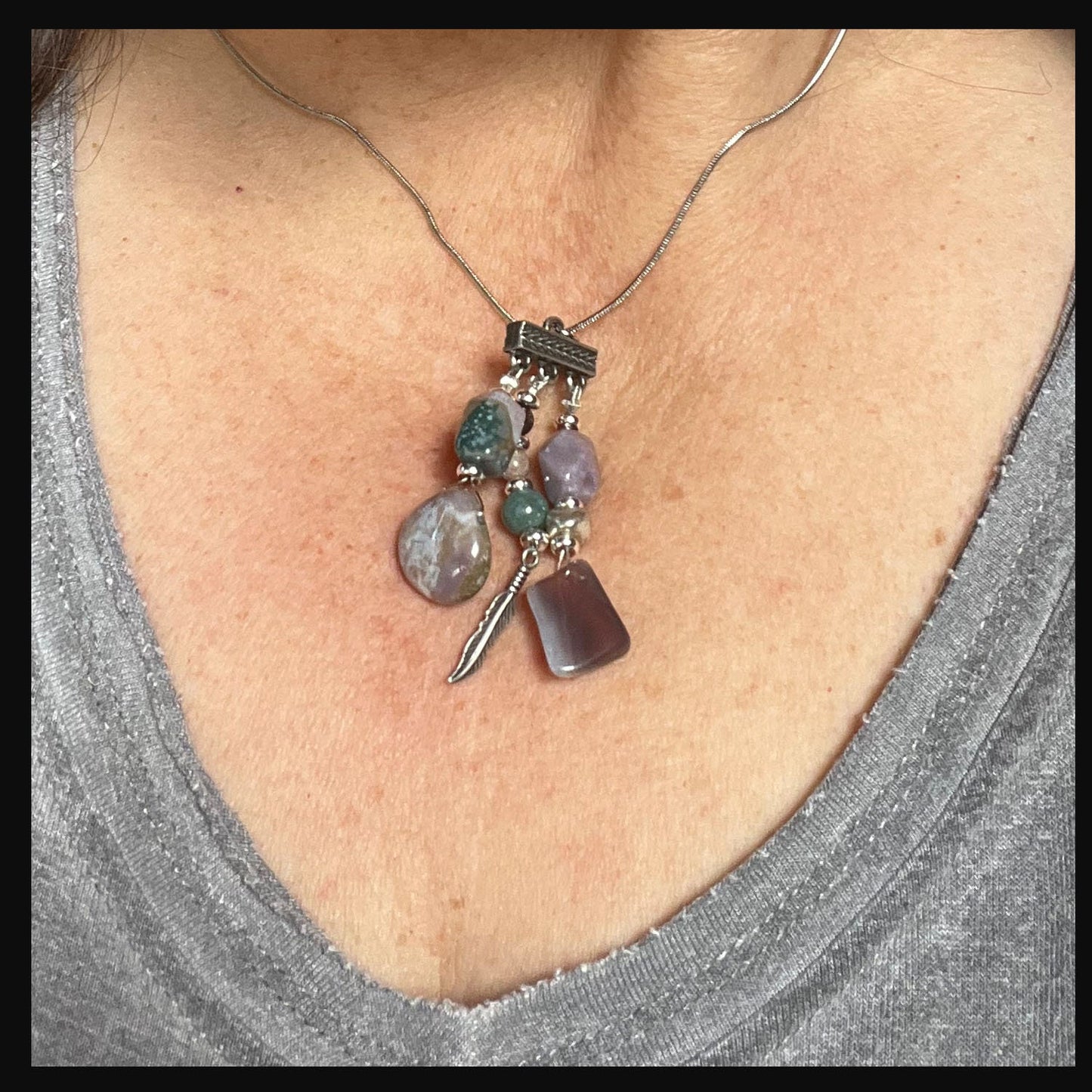 A close-up of a person wearing the Fancy Jasper Feather Pendant Necklace: Artisan Zen Jewelry, showcasing gemstone pendants in purple, green, and translucent hues against a light gray shirt.