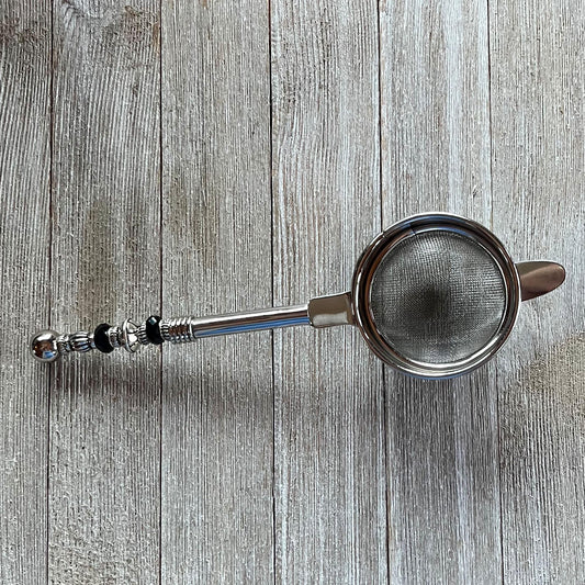 The Beaded Tea Strainer – Handcrafted Customizable Handle (9” Length), adorned with black and silver accents, rests elegantly on a light wooden surface—an ideal gift for tea lovers.