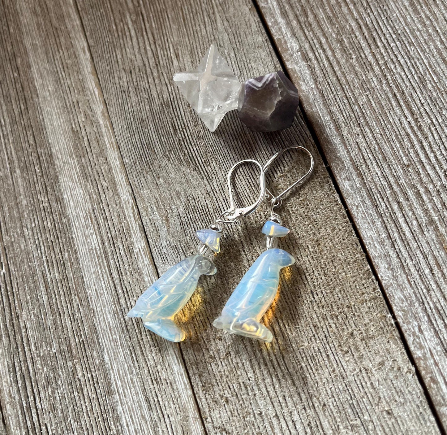 Handcrafted Opalite Penguin Earrings with sterling silver lever backs rest on rustic wooden planks, accompanied by a clear star-shaped crystal and a faceted purple stone.