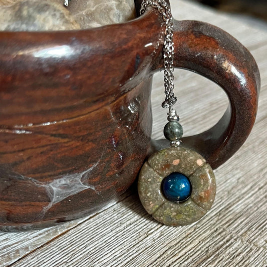 The One of a Kind Jasper Mood Pendant, featuring a color-changing bead on an 18” silver chain, hangs from the handle of a brown ceramic mug resting on a wooden surface.