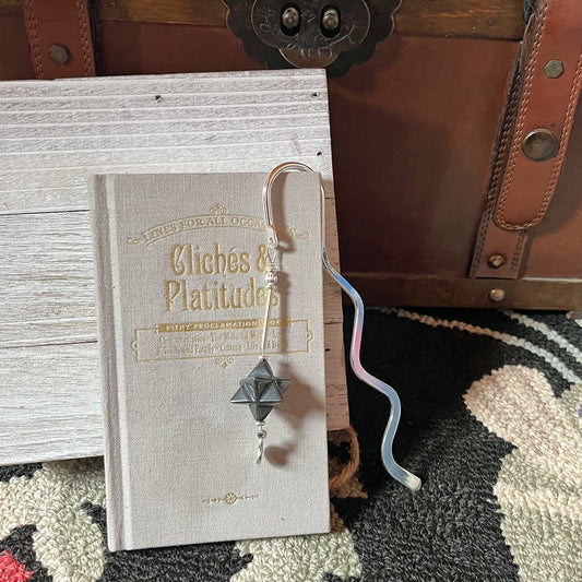 A gray book titled Cliches & Platitudes rests against a white wooden board and brown trunk. A Hematite Merkaba Bookmark: Sacred Geometry Gift for Bookworms and Readers hangs from the book, set on a patterned rug.