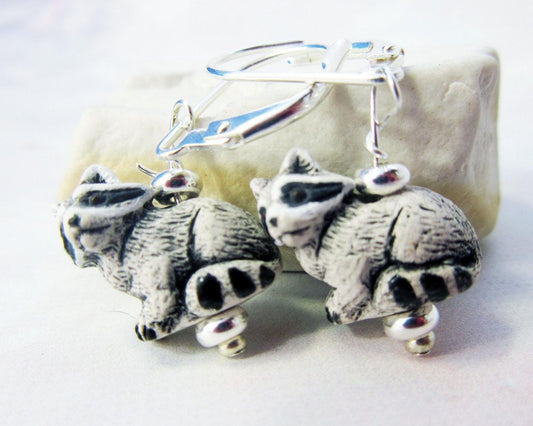 A pair of Raccoon Earrings featuring handpainted garbage panda charms with silver lever backs—perfect as a nature lover gift—displayed in front of a pale stone background.