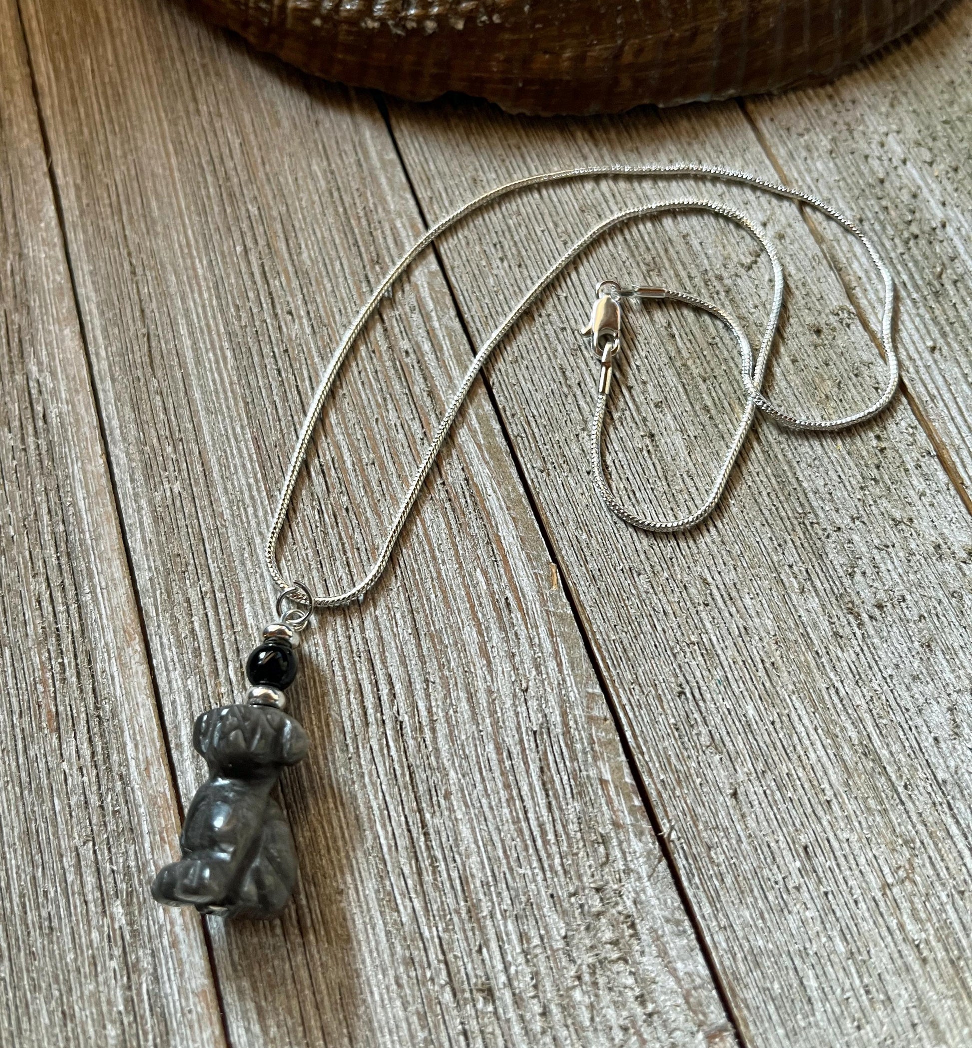 The Carved Obsidian Dog Necklace - Color Change Bead, Pet Memorial features a small dog pendant and a color-changing bead, displayed on a rustic wooden surface.