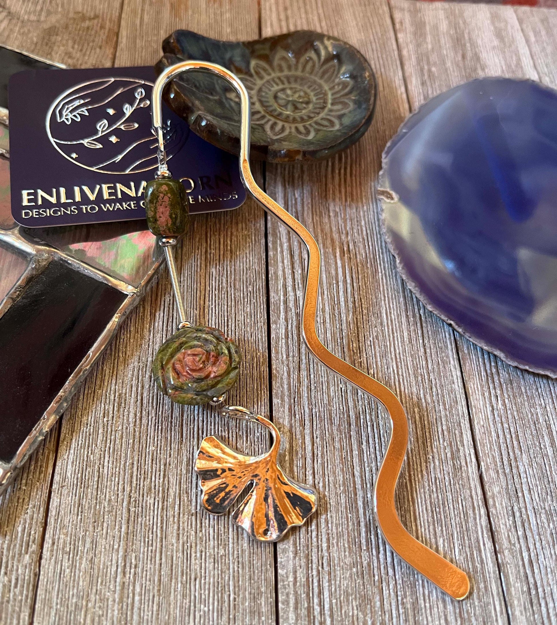 The Unakite Flower Metal Bookmark: Zen Ginko Leaf Design features green and rose-like beads on a wavy metal shape, displayed on rustic wood amid stained glass and ceramic dishes—an ideal reader gift.
