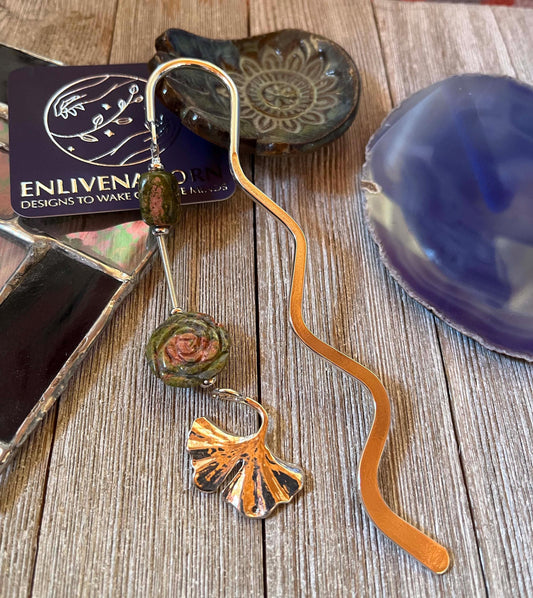 The Unakite Flower Metal Bookmark: Zen Ginko Leaf Design features green and rose-like beads on a wavy metal shape, displayed on rustic wood amid stained glass and ceramic dishes—an ideal reader gift.