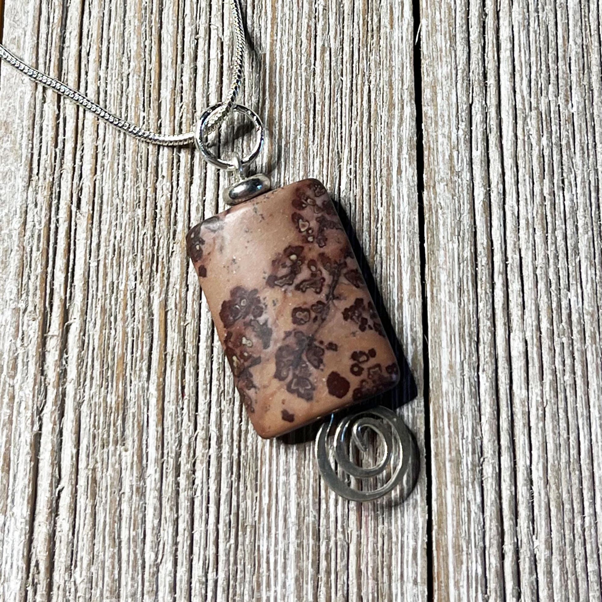 The Crazy Horse Jasper Necklace: Minimalist Zen Pendant, handcrafted in the USA, features a brown stone with dark patterns in a silver wire setting with a spiral accent and hangs from a silver chain on rustic wood.