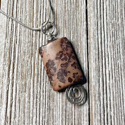 The Crazy Horse Jasper Necklace: Minimalist Zen Pendant, handcrafted in the USA, features a brown stone with dark patterns in a silver wire setting with a spiral accent and hangs from a silver chain on rustic wood.
