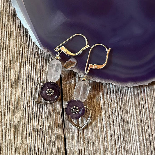 Violet Flower Earrings: Spring Flower Jewelry featuring silver hooks, clear oval beads, dark purple flower beads, and silver heart charms—a unique handmade gift or spiritual gift for wisdom. Shown on wood near a purple agate slice.