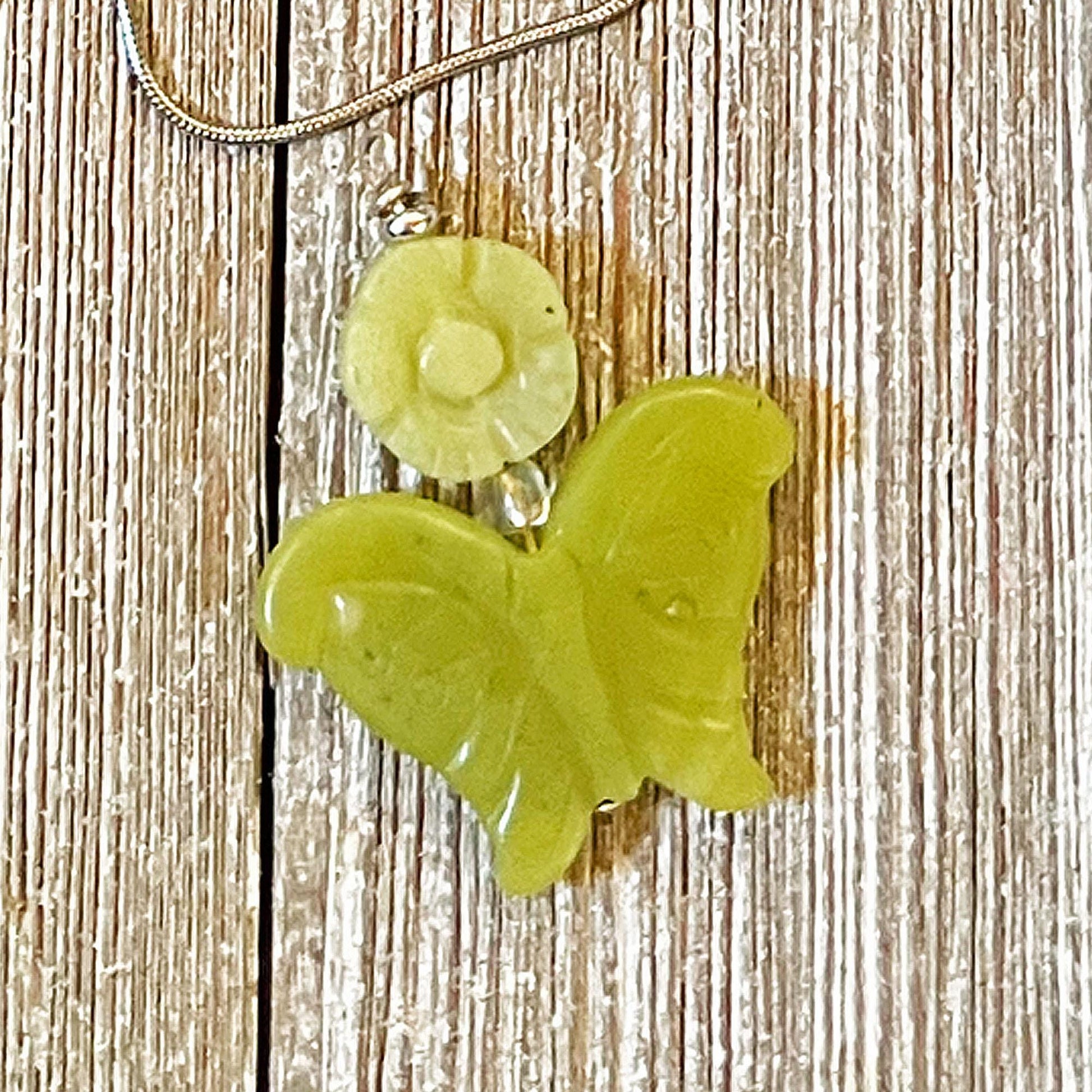 The Carved Jadeite Butterfly Pendant with Flower – Spring Nature Jewelry features a green butterfly pendant on a silver chain, beautifully showcased on textured wood.