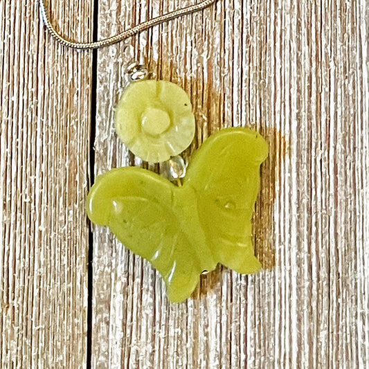 The Carved Jadeite Butterfly Pendant with Flower – Spring Nature Jewelry features a green butterfly pendant on a silver chain, beautifully showcased on textured wood.