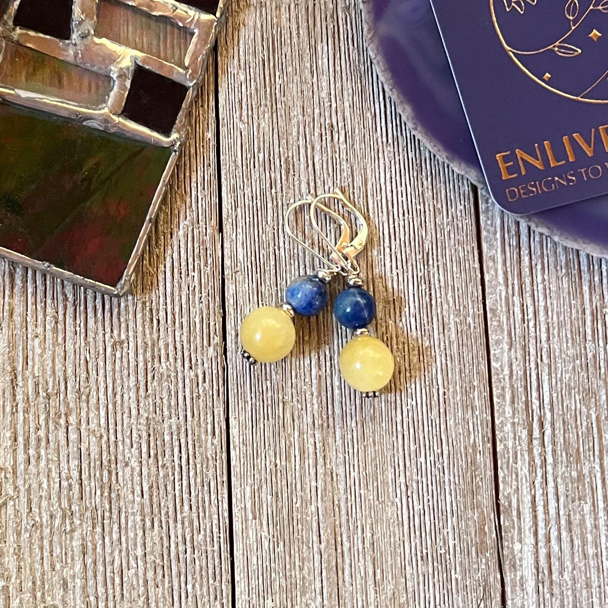 Handmade Citrine Sodalite Spring Earrings: Geometric Circles Spheres, featuring yellow and blue beads, are shown on a wooden surface with a stained glass piece and an ENLIVEN DESIGNS TO card. Ideal for lovers of unique spring or geometric earrings.