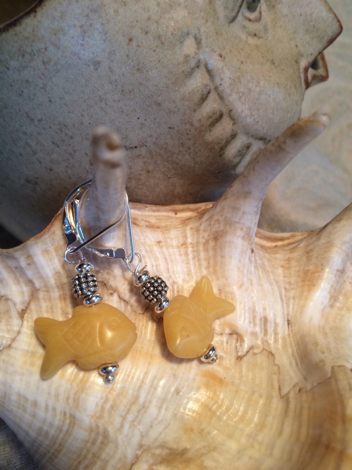 Close-up of Handcrafted Fish Earrings – Animal Jewelry, Ocean Lover Gift in yellow with silver beads, hanging from a spiral seashell; a ceramic fish sculpture appears in the background—ideal for unique ocean jewelry fans.