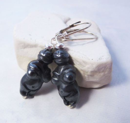 The Carved Sodalite Venus Earrings – High Priestess Jewelry, Zen Gift feature small black Sodalite figures resembling Venus pendants with silver hooks, beautifully displayed on a light textured background.