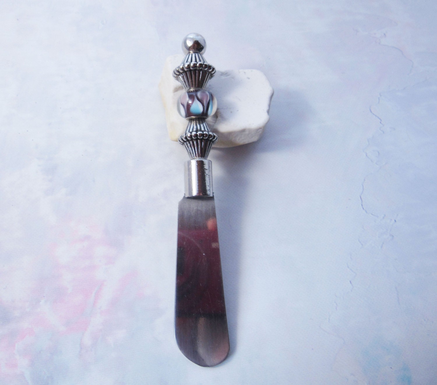 The Handcrafted Dip Spreader Knife – Glass Beads, Modern Entertaining features a silver-detailed glass bead handle and rests on white stone against a pastel background—an ideal accent for stylish gatherings.